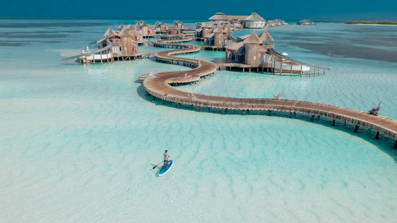 Aerial view of Soneva Maldives holiday resort with thatched-roof bungalows on stilts connected by winding wooden walkways over clear turquoise water. A person is seen paddleboarding in the shallow water, surrounded by the beautiful, serene seascape.