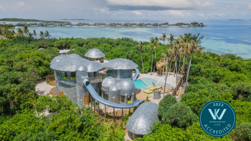 A luxurious, dome-shaped Maldives resort surrounded by lush greenery with a water slide extending from one of the domes into a pool. The Soneva resort overlooks a tropical beach with overwater bungalows in the distance. A "2023 Worldwide Kids Accredited" badge is present in the image.