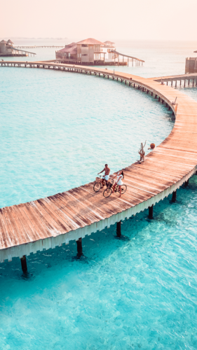 Aerial view of two people riding bicycles on a curved wooden boardwalk over clear turquoise water. The boardwalk leads to overwater bungalows in a serene, tropical setting, enhanced by Soneva Exclusive Offers. The horizon is hazy with a soft, pastel sky.