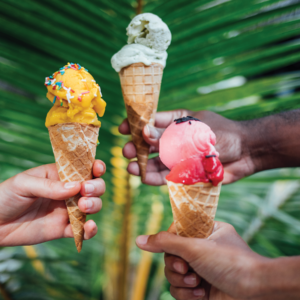 Three hands holding Soneva Maldives Luxury ice cream cones in front of a green leafy background. The left hand holds a cone with yellow ice cream and colored sprinkles, the middle hand holds a cone with green ice cream, and the right hand holds a cone with pink ice cream.