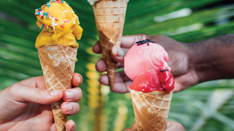 Three hands holding Soneva Maldives Luxury ice cream cones in front of a green leafy background. The left hand holds a cone with yellow ice cream and colored sprinkles, the middle hand holds a cone with green ice cream, and the right hand holds a cone with pink ice cream.