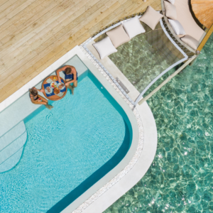 Aerial view of two people relaxing in a pool next to a wooden deck at Soneva Fushi. The pool has a round table with drinks and snacks. Adjacent to the pool, there is a netted hammock with a cozy setup including cushions. The clear ocean water and stunning Maldives scenery enhance the serene dining experience.