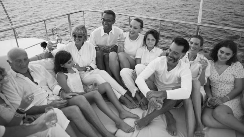 A group of people, including adults and children, are sitting closely together on a luxury yacht in the Maldives. They are all smiling and appear relaxed and happy, enjoying a moment by the water. The image is in black and white, capturing a casual, joyful scene.