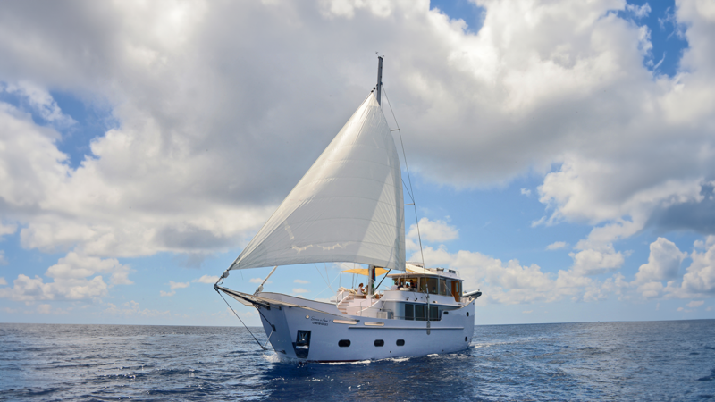 A white yacht with sails fully extended moves across a calm ocean under a partly cloudy sky. Several people are on the deck, enjoying the sunny weather of their Maldives luxury yacht holiday. The boat creates a small wake in the deep blue water.