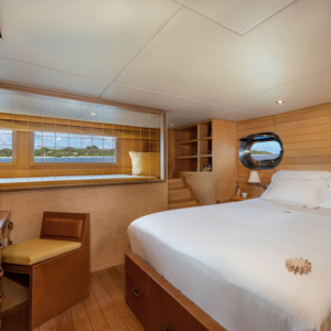 A cozy bedroom on a Maldives luxury yacht holiday features a neatly made bed with white linens, wooden furnishings, and ample storage. A unique porthole window and a large picture window offer scenic views of the water. A small desk with a chair is placed near the bed.