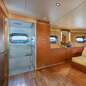 A luxurious wooden-floored room on this Maldives luxury yacht holiday features a modern bathroom with a glass door on the left, complete with a shower, porthole window, and toiletries. The right side includes cabinets, a countertop with a sink, a large mirror, and another porthole window above a yellow sofa.