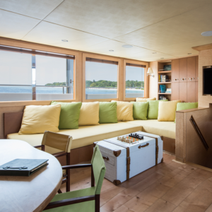 A cozy boat cabin on your Maldives luxury yacht holiday features a large U-shaped couch with yellow and green cushions. A vintage white trunk serves as a coffee table, supporting a board game. Large windows provide views of the serene sea and a distant island. A round wooden table and chair are in the foreground.