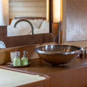 A bathroom vanity area reminiscent of a Maldives luxury yacht holiday, featuring a stylish metallic sink, a textured mirror frame, and a modern faucet. On the counter are green soap and lotion dispensers on a tray, along with a small cup. Warm lighting illuminates the cozy and elegant space.