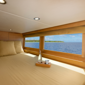 A cozy bed with beige linens inside a luxury yacht features a tray with a glass bottle and cup. Large windows offer a scenic view of the Maldives&#039; ocean and a distant tropical island under a clear blue sky. Recessed lighting illuminates the interior, enhancing your holiday experience.