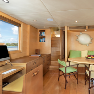 This image shows a cozy, well-lit room on a Maldives luxury yacht holiday with wooden floors and furniture. It has a desk with an iMac, shelves with books, a round table with green cushions, chairs, and a wall-hung decorative ship wheel. A staircase leads to an upper level.