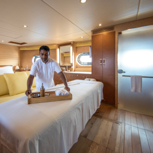 A spa therapist arranges a tray with rolled towels and massage oil on a massage table in a serene, well-lit room with wooden floors and soothing decor. The cozy setting includes plush yellow cushions, large windows, and reminders of a Maldives luxury yacht holiday.