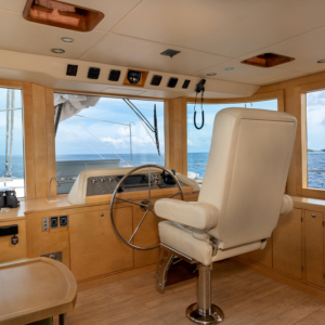 The interior helm station of a luxury yacht, perfect for a Maldives holiday, features a large white captain&#039;s chair, a wooden steering wheel, and various control panels. The view out the wide windows showcases a clear, sunny day with a calm ocean and blue sky in the background.