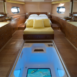 The interior of a luxurious yacht&#039;s cabin features a bed with yellow bedding and pillows, wooden cabinetry, and two circular windows. There&#039;s a glass floor panel revealing an aquarium beneath, showcasing fish swimming in the illuminated water below—a perfect setting for your Maldives Luxury Yacht Holiday.