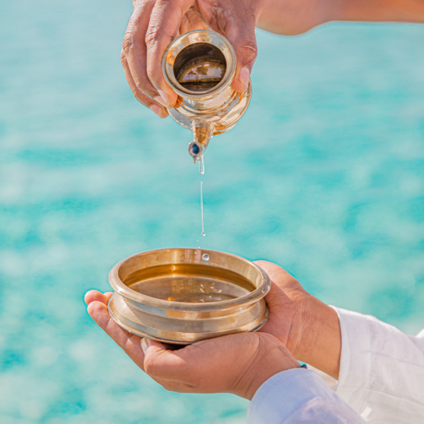 A close-up of hands against a turquoise background. One pair of hands holds a small metal pot, from which clear liquid is being poured into a round metal container held by another pair of hands. Captured mid-drip, the scene radiates the precision and exclusivity found in Soneva Exclusive Offers.