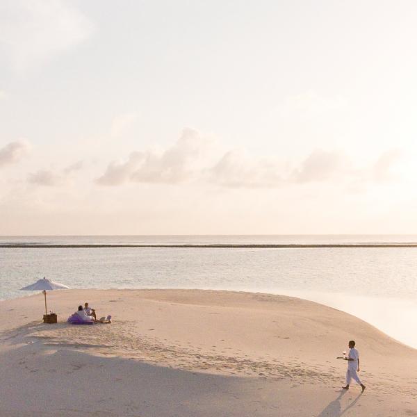 A peaceful beach scene at sunset shows two people relaxing under a white umbrella on a small sandy island. Another person, dressed in white, walks toward them carrying a tray. The sky is pastel-colored with soft clouds, and the calm sea surrounds the tiny island—truly embodying Soneva Exclusive Offers.