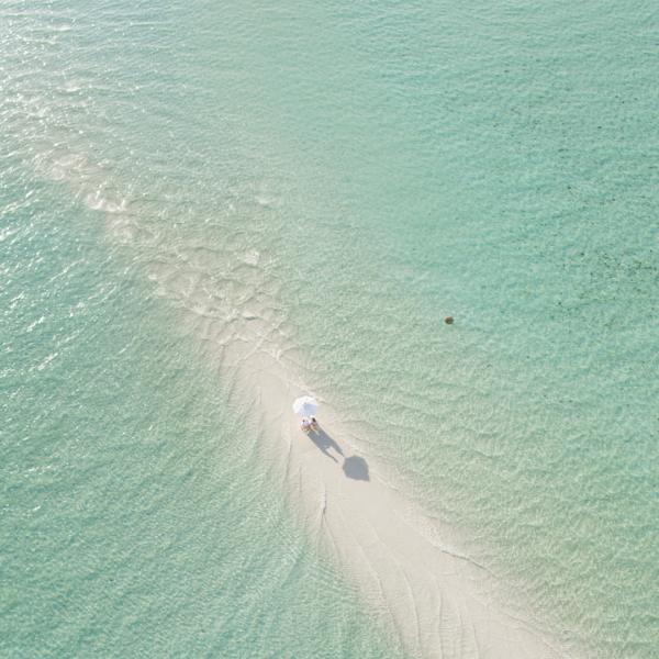 Aerial view of a person with an umbrella standing on a narrow strip of sand surrounded by clear, turquoise water. The scene gives a sense of tranquility and isolation, perfectly capturing the essence of Soneva Exclusive Offers. The person’s shadow and the umbrella are clearly visible on the sand.