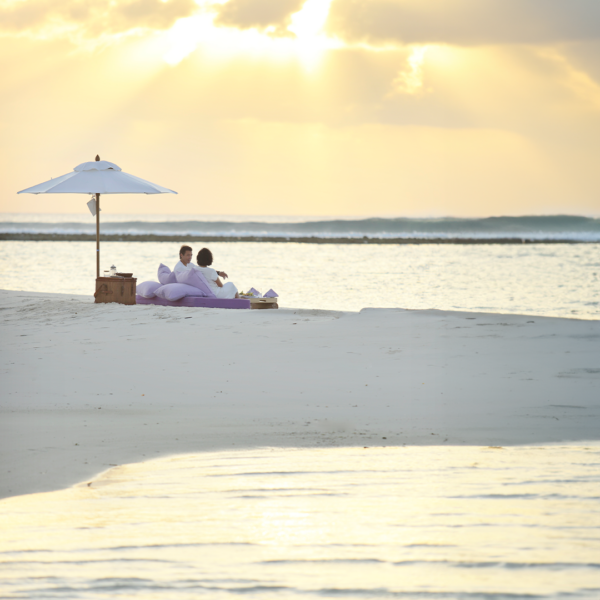 Two people sit closely together on cushions under a white beach umbrella at the edge of a tranquil beach. The sun is setting, casting a golden glow over the water and creating a serene, romantic atmosphere—a moment made even more special with Soneva Exclusive Offers.