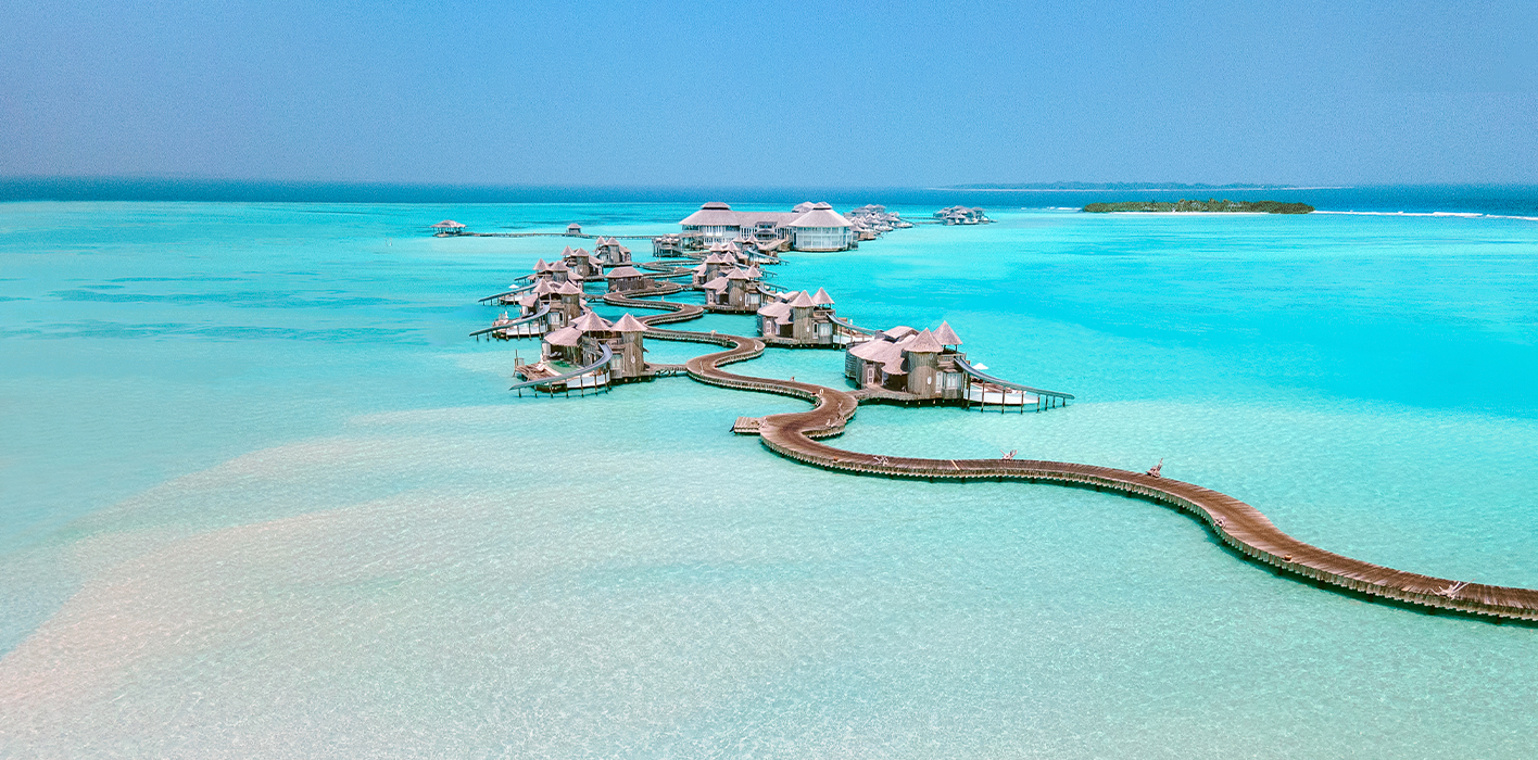 Aerial view of luxury overwater villas connected by winding wooden walkways in a turquoise lagoon at Soneva Maldives Resort. The structures are surrounded by crystal clear water extending to the horizon, with a small lush island in the background. The sky is clear and blue.