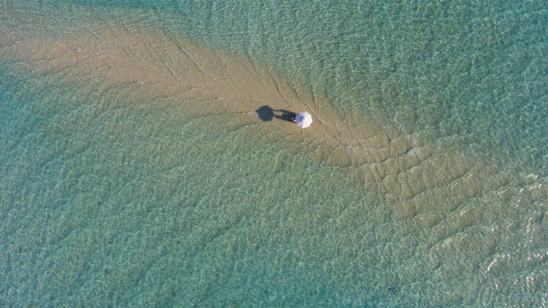 Aerial view of a solitary beach umbrella casting a shadow on a sandy strip surrounded by clear, shallow turquoise water. The isolated setting at this Maldives Luxury Resort evokes a sense of peaceful seclusion and tranquility.