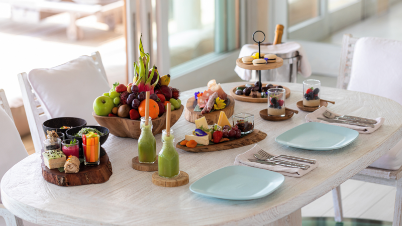 A beautifully set table with a variety of fruits, vegetables, cheeses, desserts, and drinks reminiscent of a Maldives luxury resort experience. The table features plates, cutlery, and glasses ready for dining. The bright, airy room with large windows feels like a touch of Soneva elegance.