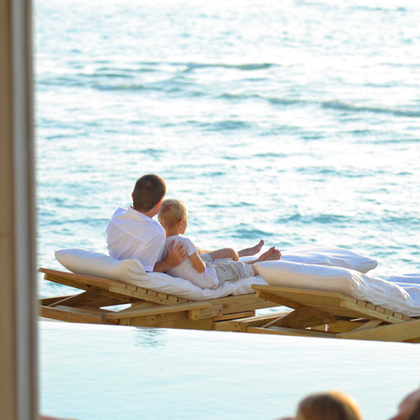 A man and a child are lying on a lounge chair over a body of water, looking at the ocean. In the foreground, another person is also reclining on a lounge chair with their back turned. The serene scene features clear, calm water and a bright sky, perfect for enjoying Soneva Exclusive Offers.