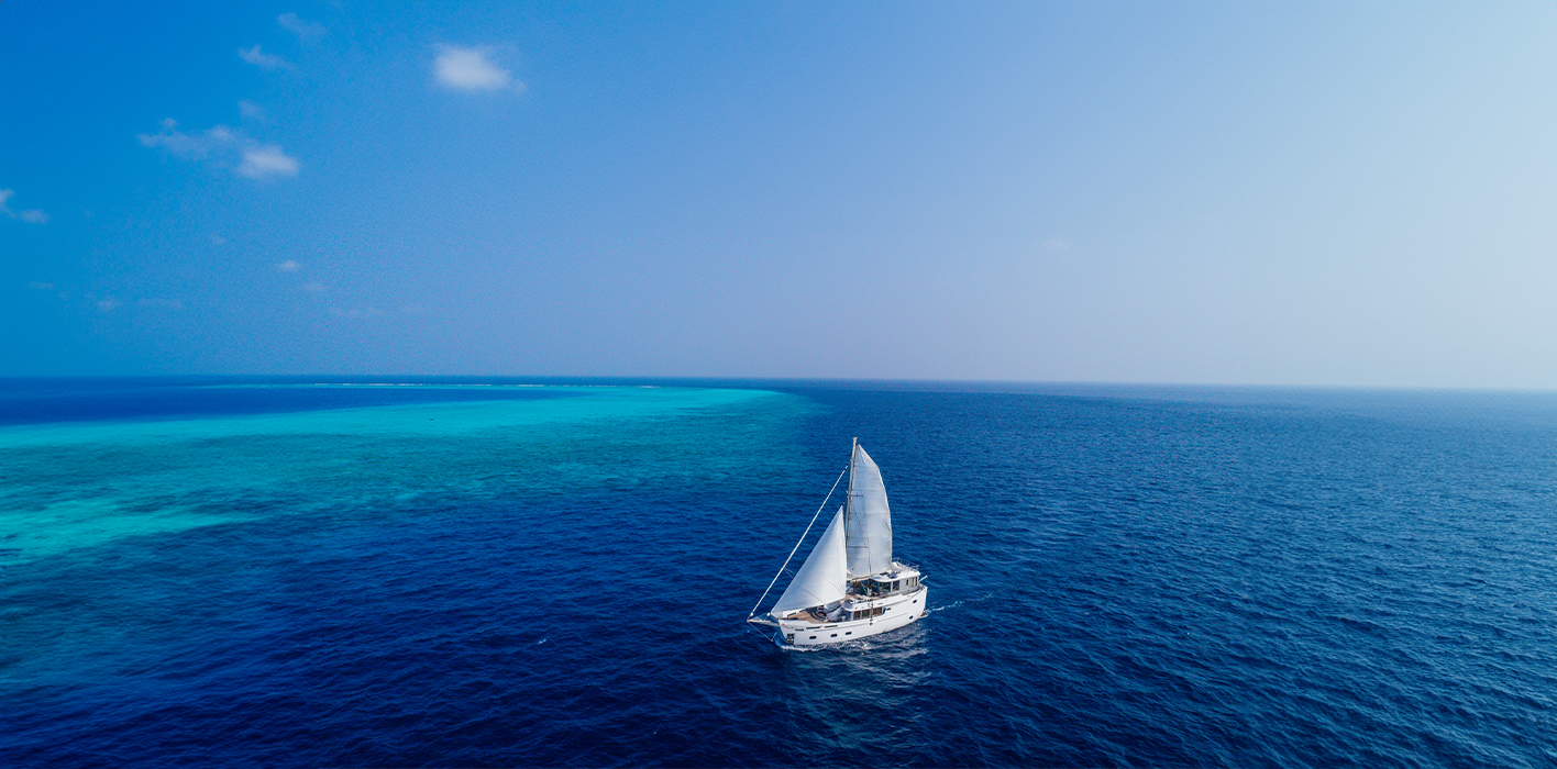 A luxurious sailboat with white sails glides across a vast, deep blue ocean under a clear sky, offering the epitome of a Maldives holiday. The horizon shows gradients of water shades, transitioning from turquoise near a visible reef to darker blue further out. Sparse clouds dot the sky.