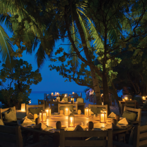 A romantic, candlelit dinner setup on a beach during the evening at Soneva Fushi. Round wooden tables and comfortable chairs are arranged under palm trees, with lanterns illuminating the area. The dark blue sky and the sea create a tranquil backdrop for this exquisite Maldives dining experience.