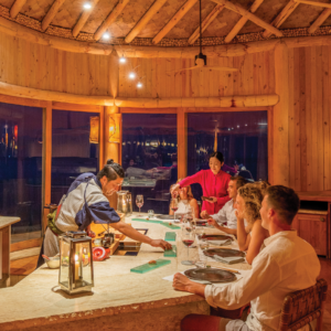 A chef in traditional attire prepares food at a circular sushi bar in a warmly lit, rustic, wooden restaurant. Three patrons sit around the bar with wine glasses, while a server pours wine for a guest. The atmosphere is as cozy and inviting as Maldives dining at Soneva Fushi.