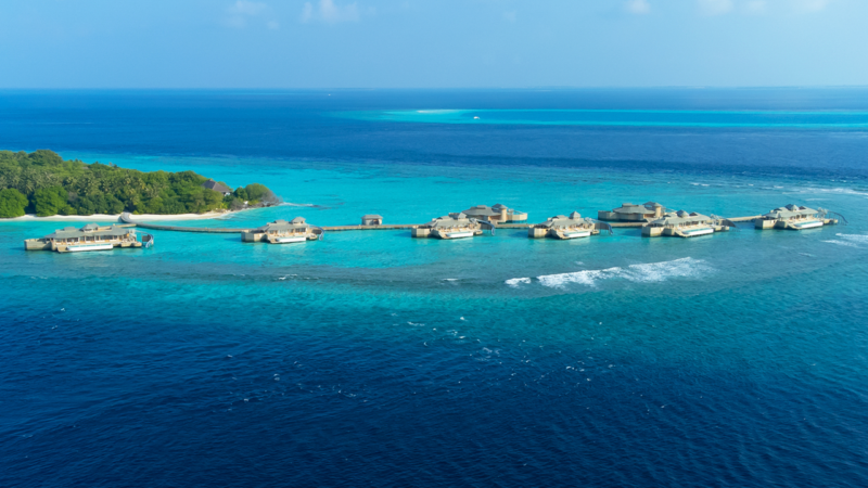 Вид с воздуха на Soneva Fushi Villas, роскошный курорт, построенный над бирюзовыми водами на небольшом острове на Мальдивах, окруженном глубоким синим океаном. Курортные строения соединены между собой пешеходными дорожками, а часть острова покрыта пышной зеленью под ярким, чистым небом.