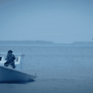 A person stands at the bow of a small, white boat in a calm, blue ocean near the Maldives. A signpost with a star-shaped marker is visible in the water beside the boat. The horizon is hazy, creating a serene and tranquil atmosphere, reminiscent of the peaceful Namoona islands.