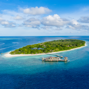 Aerial view of a tropical island surrounded by turquoise waters and white sandy beaches. The island is lush with greenery and has several buildings, including some overwater structures extending into the ocean. Experience Soneva Exclusive Offers as you relax under clear skies with scattered fluffy clouds.