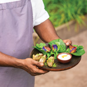 A person wearing a white shirt and gray apron holds a wooden plate with green leaves, edible flowers, stuffed shell-like food items, and a small bowl of dipping sauce. The scene evokes the lush, tranquil ambiance of Maldives dining at Soneva Fushi, with blurred greenery in the background.