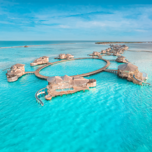 Aerial view of luxurious overwater villas in a circular layout on turquoise waters during a Soneva Maldives holiday. The wooden bungalows are connected by a curved boardwalk, surrounded by clear, blue ocean under a bright blue sky. Distant land is visible on the horizon.