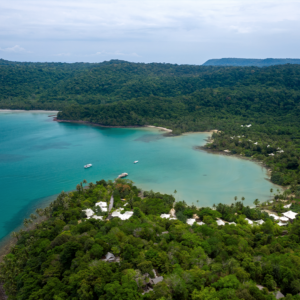 Aerial view of the tropical coastline at Soneva Kiri Resort featuring lush green foliage, a secluded beach, and turquoise waters. There are a few white-roofed buildings nestled among the trees. The sky is partly cloudy, and the water has a teal hue blending into deeper blue shades.