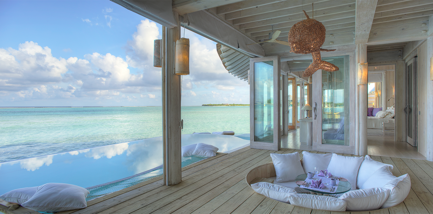 Luxurious beachfront villa at the Maldives Luxury Resort Soneva overlooking the ocean with an infinity pool. Features include an outdoor sunken seating area with white cushions, wooden decking, and glass doors. Nautical decor and plush accents enhance the relaxed, upscale ambiance.