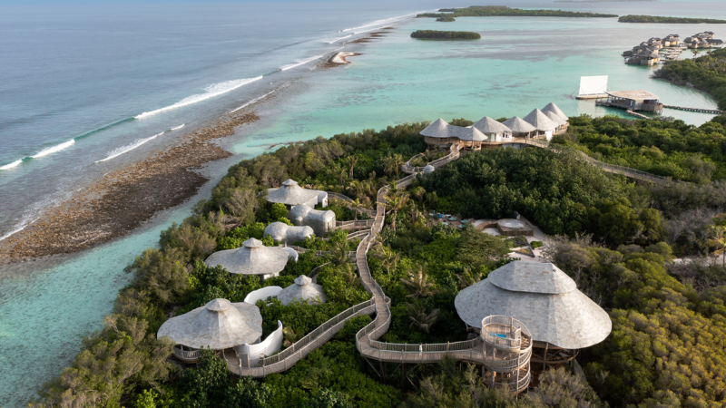 Aerial view of the luxurious Soneva resort in the Maldives, featuring several thatched-roof villas interconnected by elevated wooden walkways. Surrounded by lush greenery, it overlooks a vibrant coral reef and turquoise ocean waters, with more villas visible in the distance.