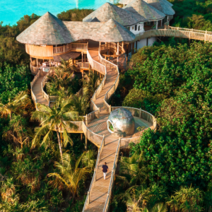 Aerial view of a tropical resort featuring wooden pathways weaving through lush green foliage, leading to elevated round villas overlooking a turquoise ocean. In the distance, additional villas on stilts over the water. A person walks along the pathway, embodying the Maldives exclusive experience at Soneva.