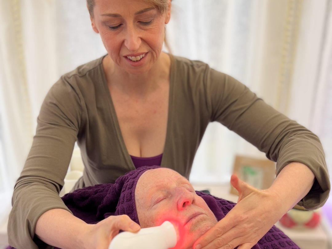 A woman is giving a facial treatment to another woman using a handheld device emitting red light. The client is lying down with a towel covering her hair, and both are in a softly lit room. The atmosphere appears calm and focused.