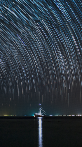 A long-exposure photo capturing a boat illuminated on the calm sea at Soneva in the Maldives under a night sky filled with dramatic star trails. The water reflects the light from the boat and the starry sky, enhancing this exclusive experience with its serene scene.