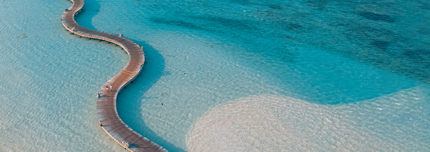 Aerial view of a long, winding wooden boardwalk stretching over clear, shallow turquoise water at the Maldives luxury resort Soneva. People can be seen walking along the boardwalk. The water transitions from light to dark shades, indicating varying depths.