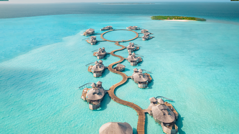 Aerial view of luxurious overwater villas at a Maldives luxury resort, Soneva, arranged in a serpentine pattern in a clear teal ocean. Each villa has a thatch roof, connected by winding wooden walkways. In the distance, a small green island sits on the turquoise horizon.