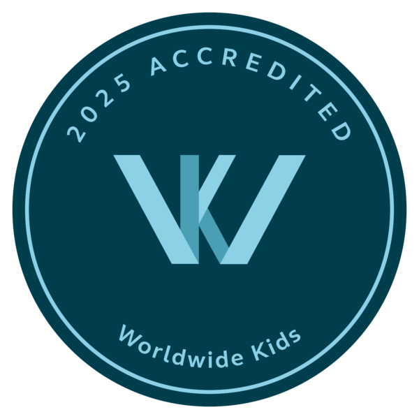 A circular emblem with the text "2025 Accredited" at the top and "Worldwide Kids" at the bottom sits proudly, reminiscent of a family resort in the Maldives. At its center, a stylized "W" and "K" intertwine gracefully in light blue on a dark teal background.